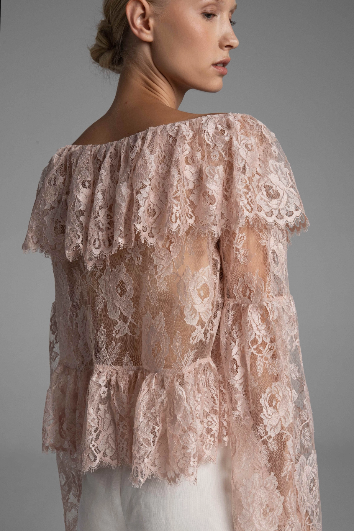 TThe back of an ethereal blouse made of French lace by Monika Adamska in a blush shade, featuring an asymmetrical hem and intricate weave.